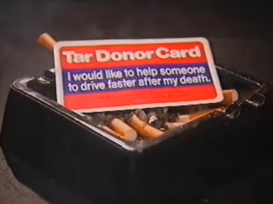 SCREENCAP: An ashtray full of fag-ends, on top of which is a card marked "Tar Donor Card. I would like to help someone to drive faster after my death."